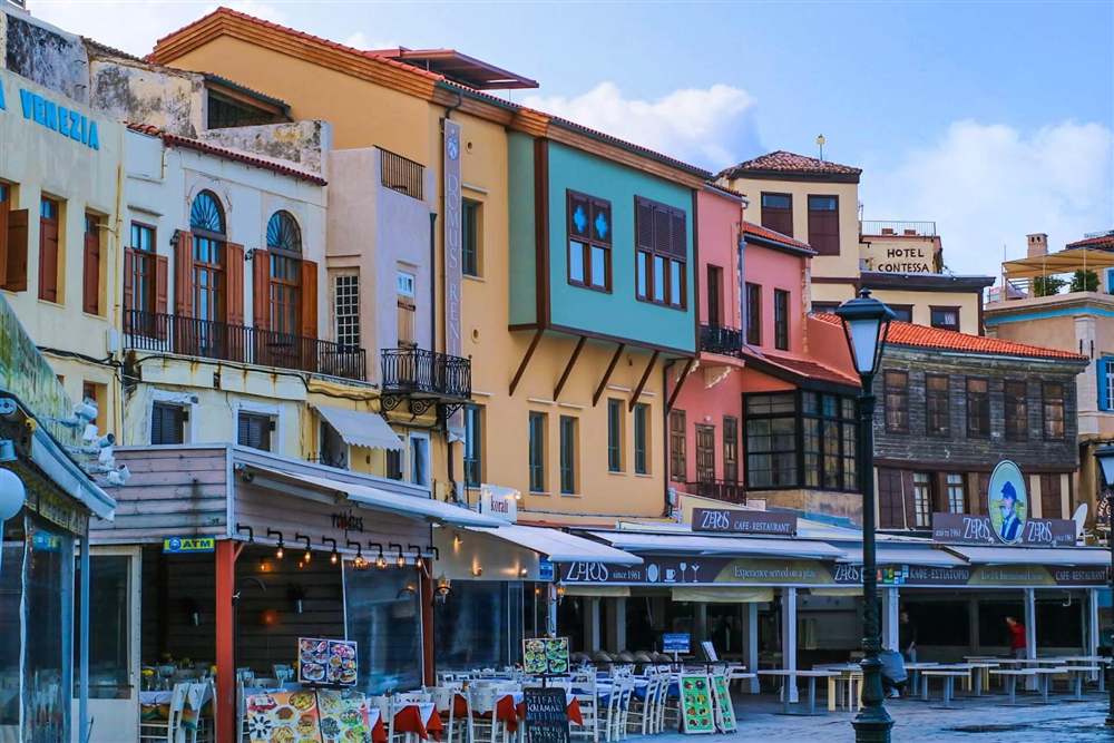 Mykonos, Santorini and Crete tour: a colourful old town street on Crete with pastel houses, wooden balconies and café tables set out under a blue sky