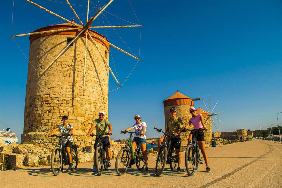 Santorini, Crete and Rhodes tour: a tall stone windmill with wooden sails and a smaller windmill on the harbour mole of Rhodes, with a few cyclists at the base under a blue sky