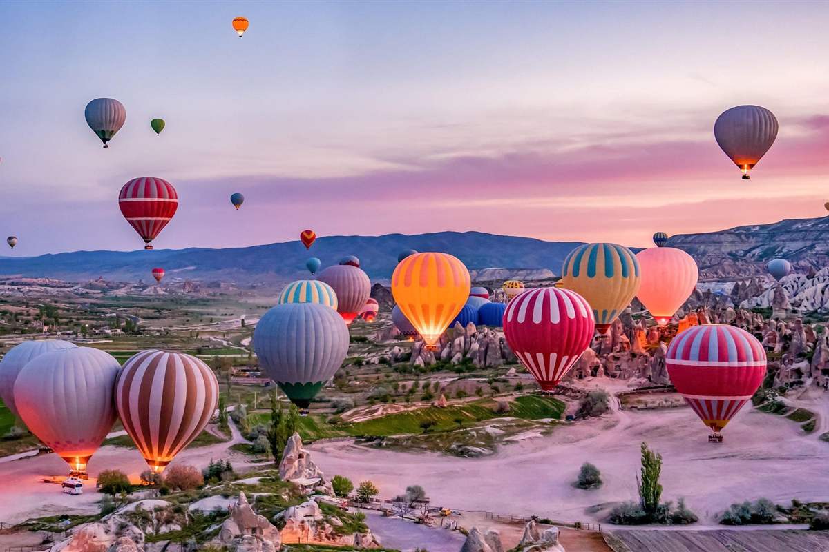 Hot air balloons rise at dawn over the rock formations and valleys of Cappadocia, with several balloons glowing from their burners and a pink and purple sky above distant mountains.