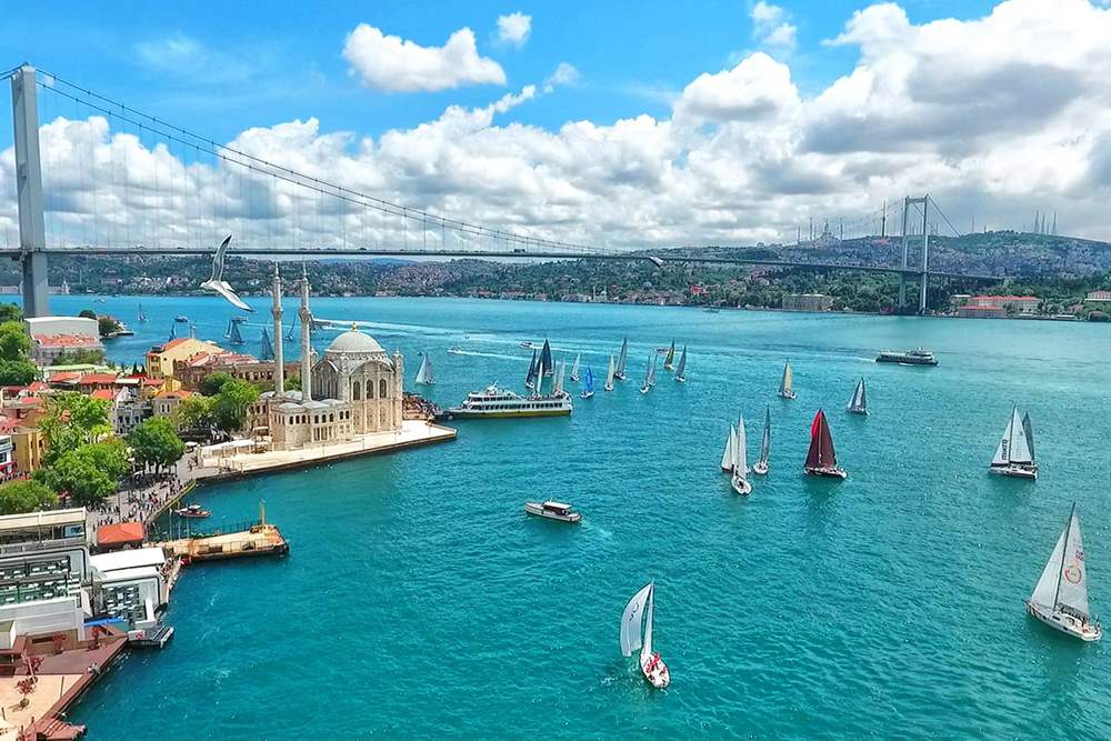 Mosque on the waterfront and a suspension bridge over a turquoise strait with sailboats and a ferry, seen from above under a blue sky with white clouds.
