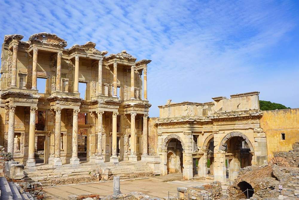 Tour in the steps of Saint Paul at an ancient two-storey stone library facade with columns and statue niches, beside a large arched gateway, under a blue sky with thin white clouds.