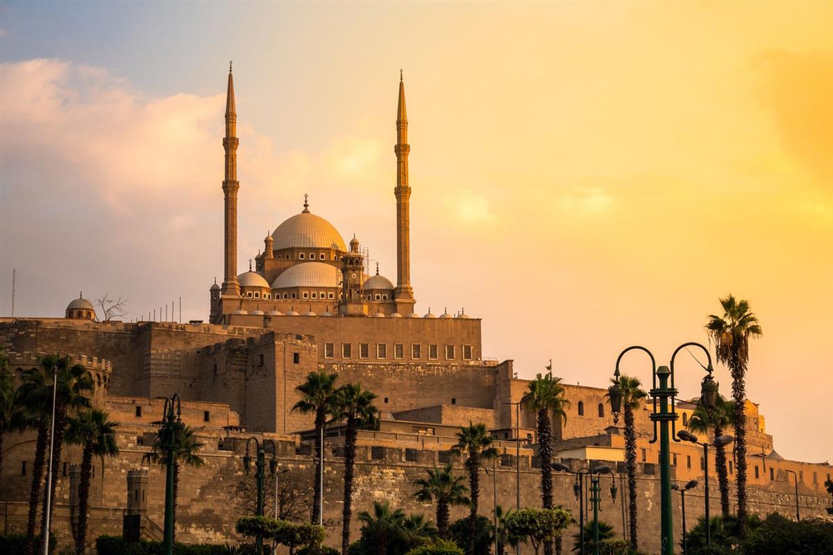 Greece and Egypt tour sights: a large domed mosque with two tall minarets rises above a stone fortress wall, with palm trees and street lamps in front under a golden sunset sky.
