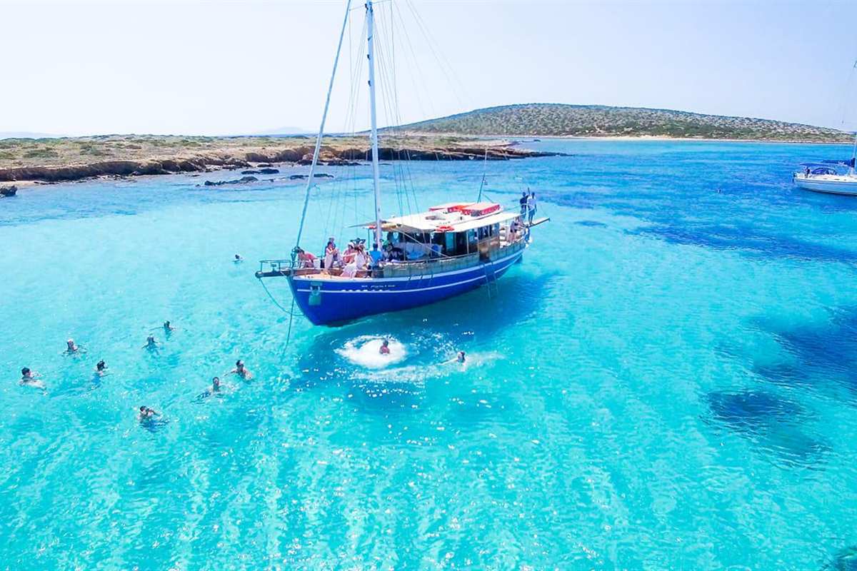 Island hopping tour: a wooden sailing boat on bright turquoise water beside a low rocky island, with swimmers around the boat and another sail in the distance