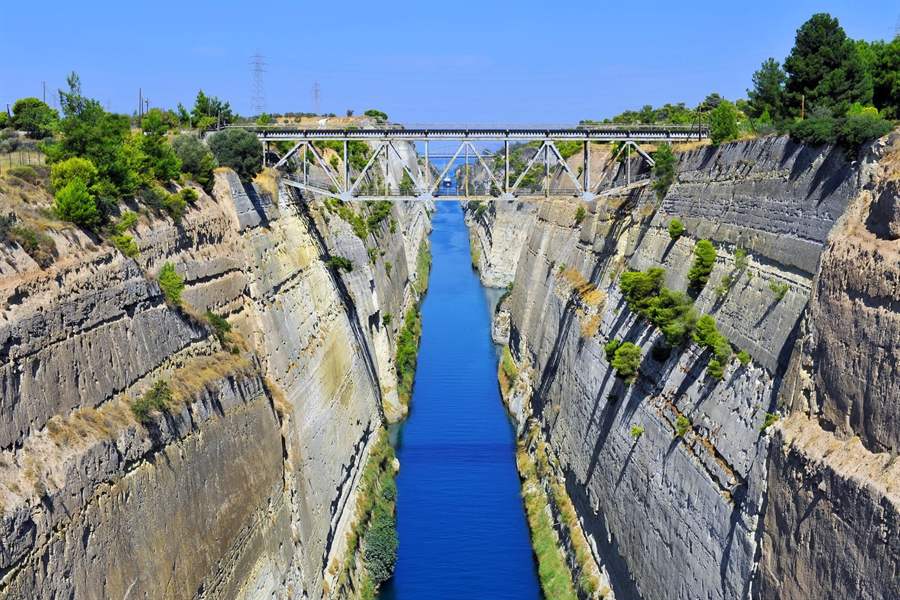 Classical tour of Greece: the narrow Corinth Canal cut deep through layered grey rock walls, blue water below and a steel truss bridge crossing high above under a clear sky