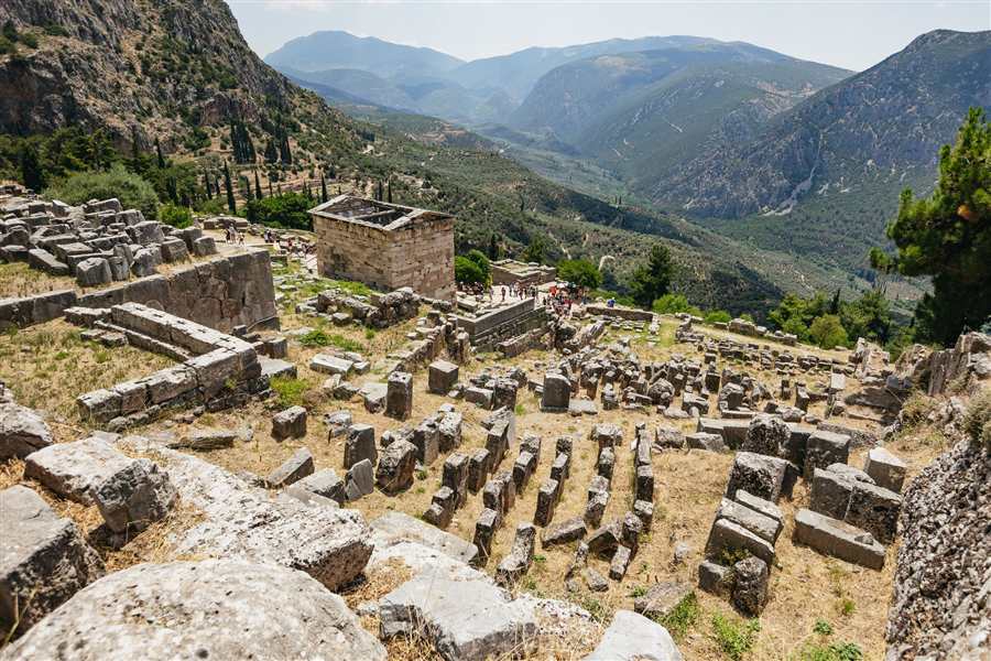 Delphi to Meteora tour: the stone Treasury of the Athenians among the ruins of the Sanctuary of Apollo at Delphi, above a deep green valley of olive groves and mountains
