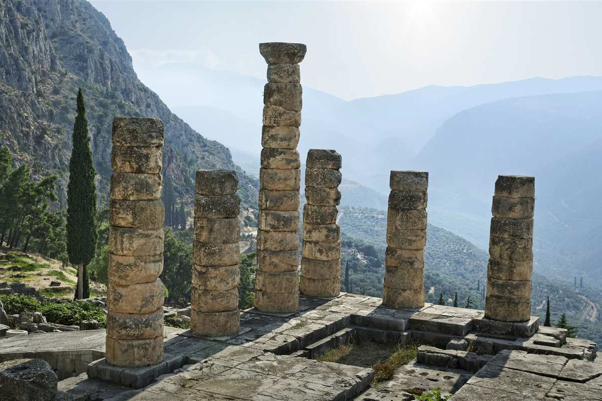 3 day Delphi Meteora tour: six ancient columns of the Temple of Apollo at Delphi stand on a stone platform, with a hazy mountain valley of olive groves and cypress trees behind them