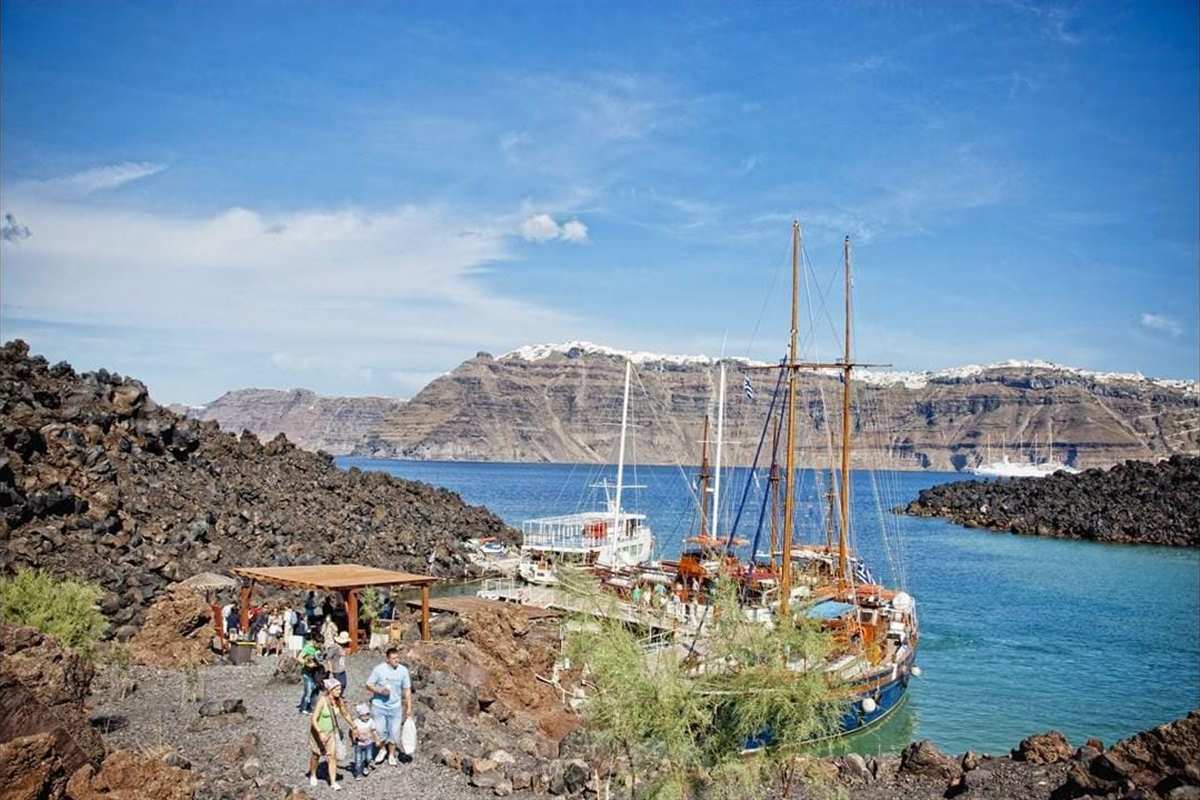 Golden Splendor Greek cruise: wooden tour boats moored in a rocky bay with a black volcanic shore, visitors on a gravel path, and the layered cliffs of a caldera across the water