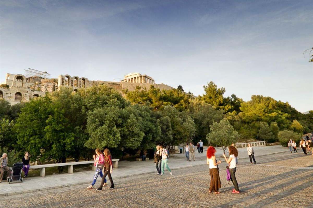 Athens stopover tour: people walking along a wide paved path under pine trees at the foot of the Acropolis, with the ancient hill and its walls above