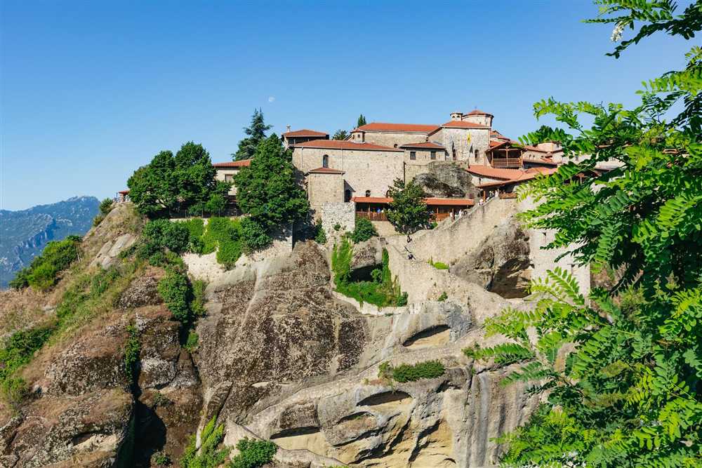 Classical tour with Meteora: a stone monastery with red-tiled roofs on top of a huge rock, stone stairs cut up the cliff, green trees around it and mountains under a clear blue sky