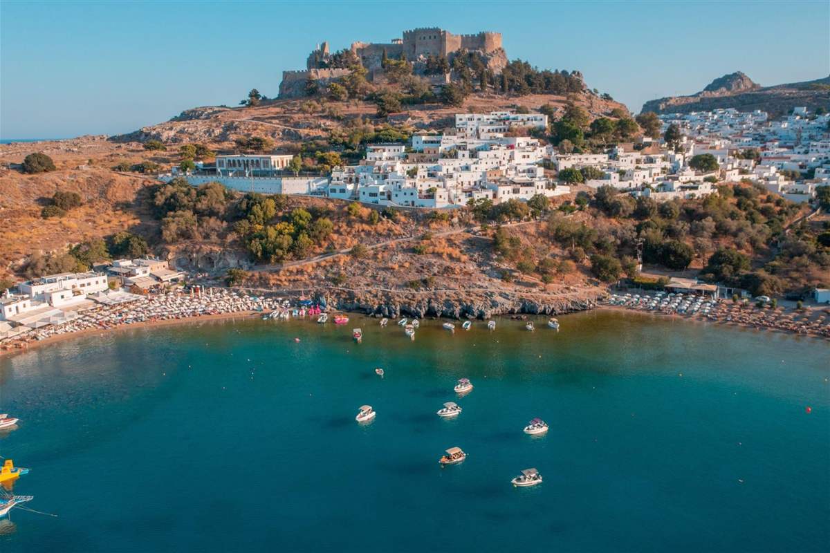 Aerial view of a curved Aegean bay with turquoise water and small boats, a white village climbing the hillside and a stone fortress on the hilltop above the beach.