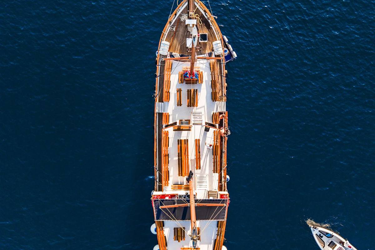 Golden Classic Greek cruise: aerial view of a wooden ship with benches on its white deck, on deep blue sea, with a small white boat at the lower right corner