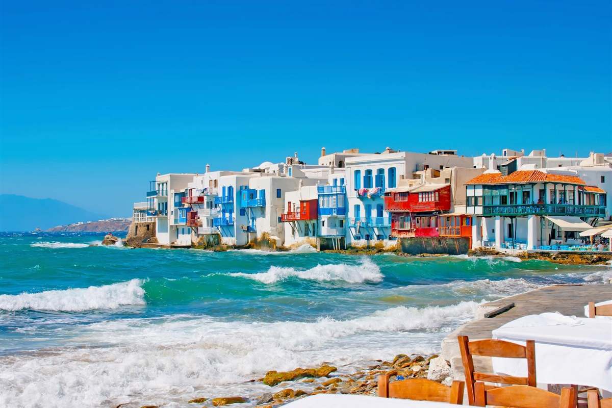 Greece Mykonos tour: white and colourful houses with balconies built right on the water at Little Venice, waves breaking on the rocks in front under a clear blue sky