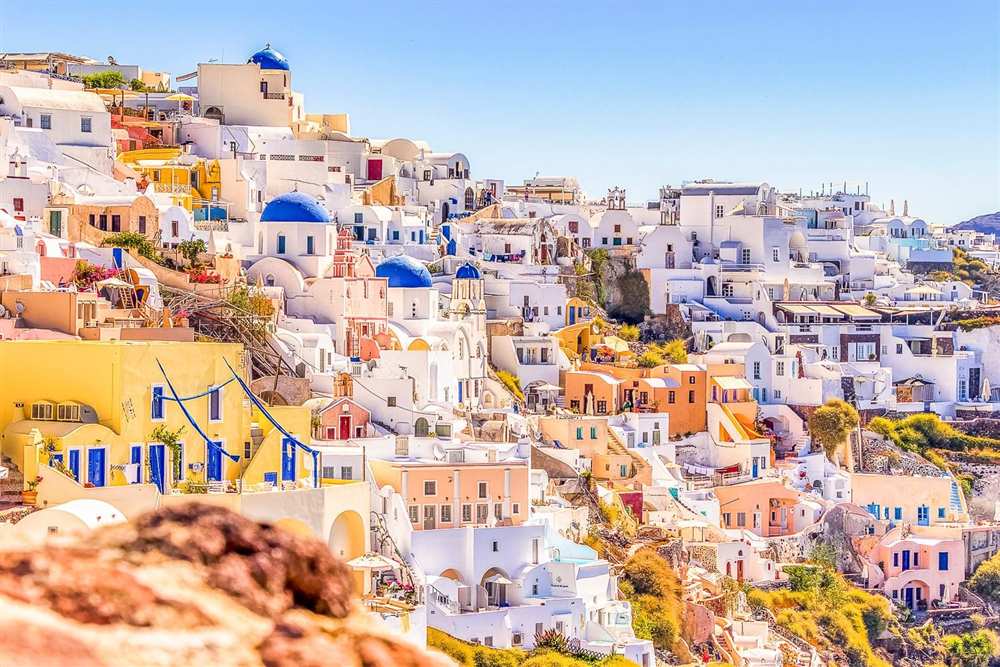 Santorini tour: white and pastel houses stacked on the hillside of Oia with several blue church domes and bell towers, under a clear blue sky