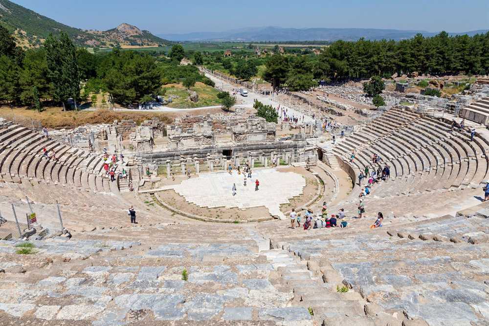 Great Theatre of Ephesus seen from the top rows, with curved stone seating, the marble stage floor, a colonnaded road leading away and green hills in the distance under a blue sky.
