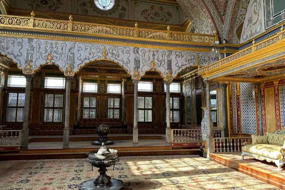 Ornate palace hall with gilded balconies, painted arches, slim marble columns, latticed windows, a silver brazier on a patterned carpet and a gold cushioned sofa in sunlight.