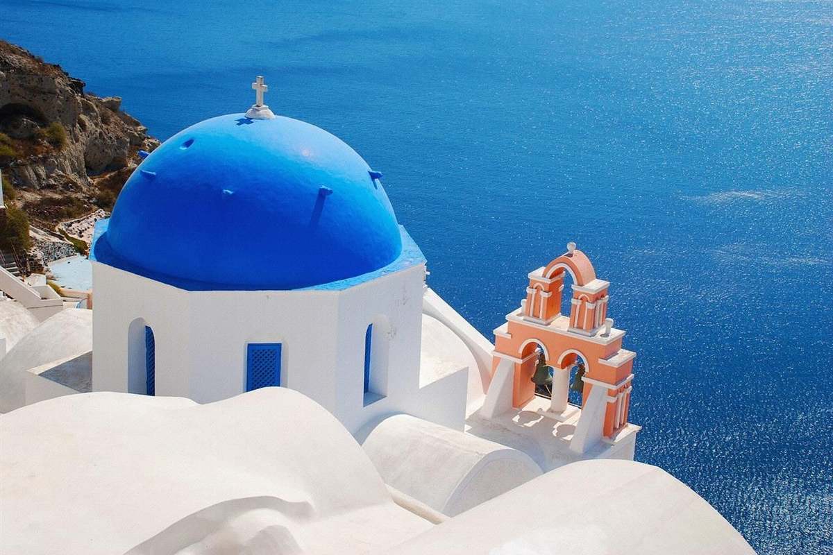 Santorini and Crete tour: a white chapel with a blue dome and a pink bell tower on the edge of the caldera, with the deep blue sea far below