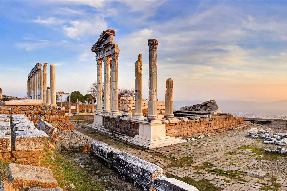 Ancient ruined temple with tall fluted stone columns and a partly standing entablature on a hilltop platform of stone paving, lit by warm sunset light under a blue and pink sky.