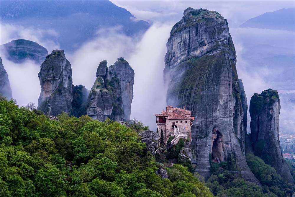 Aegean Explorer tour: tall rock pinnacles of Meteora wrapped in mist with a small monastery on a ledge below and green forest on the slopes