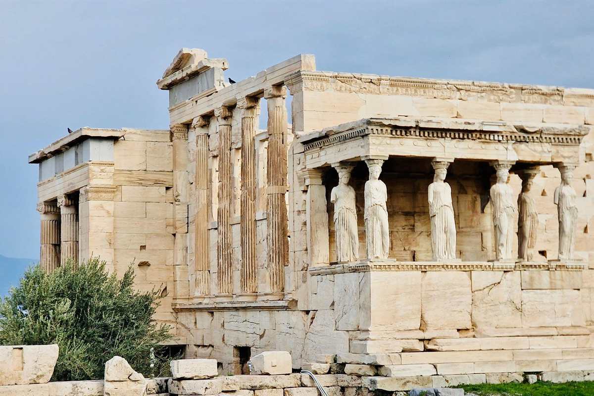 Acropolis small group tour: the Erechtheion on the Acropolis, with the Porch of the Caryatids showing six marble maiden statues on a tall stone base and fluted columns beside it, a green olive tree on the left and a grey-blue sky behind