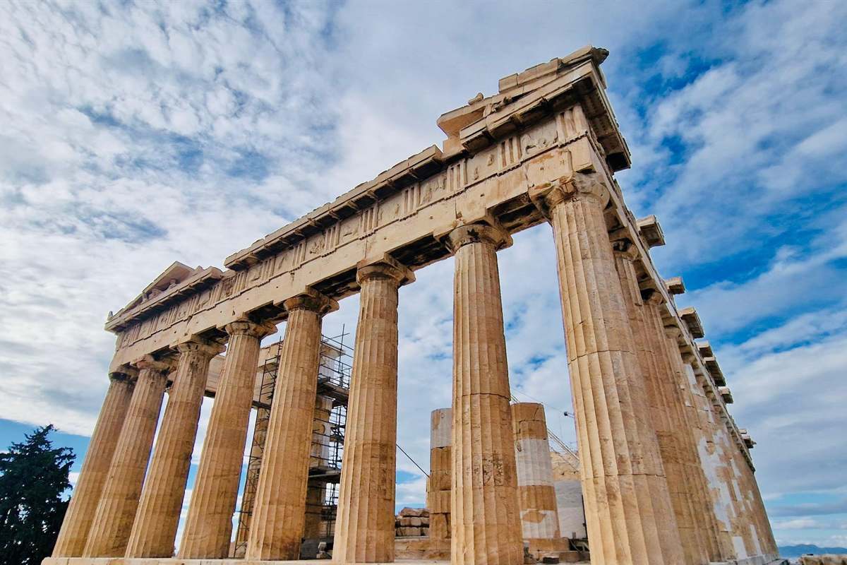 Acropolis small group tour: the Parthenon seen from a low angle at its corner, with fluted golden Doric columns, the carved frieze above them and scaffolding inside the temple, under a sky of white clouds and patches of blue