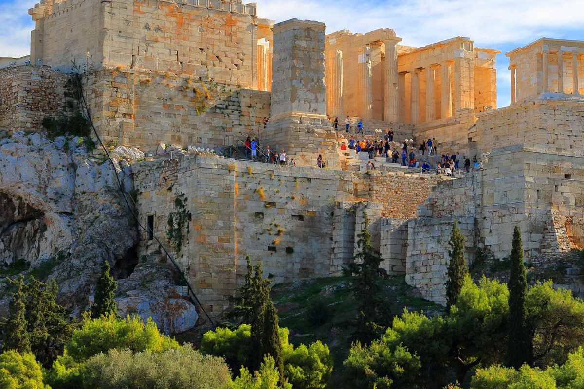 Acropolis ticket with audio guide: the sunlit marble Propylaea and the Temple of Athena Nike on top of the Acropolis rock, with green pines below and a clear blue sky