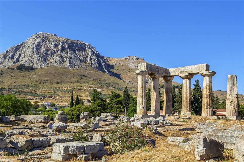 Aegean escape tour: the standing Doric columns of the Temple of Apollo at Ancient Corinth with the rocky peak of Acrocorinth behind under a clear blue sky