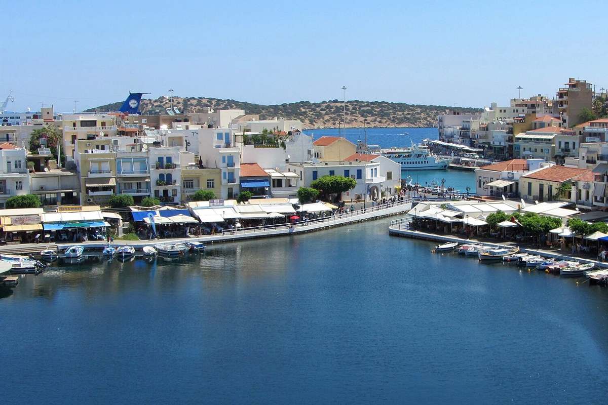 Agios Nikolaos semi-submarine: Lake Voulismeni in the middle of Agios Nikolaos town, with white and ochre houses, cafe awnings and small boats along the quay, a narrow channel leading to the harbour with a white passenger boat and a green hill behind, under a clear blue sky