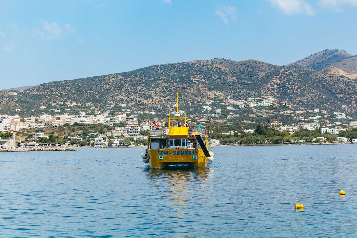 Agios Nikolaos semi-submarine: a yellow catamaran-style semi-submarine boat with passengers on its upper deck sailing on calm blue water, with white houses along the shore and olive-green hills behind under a blue sky