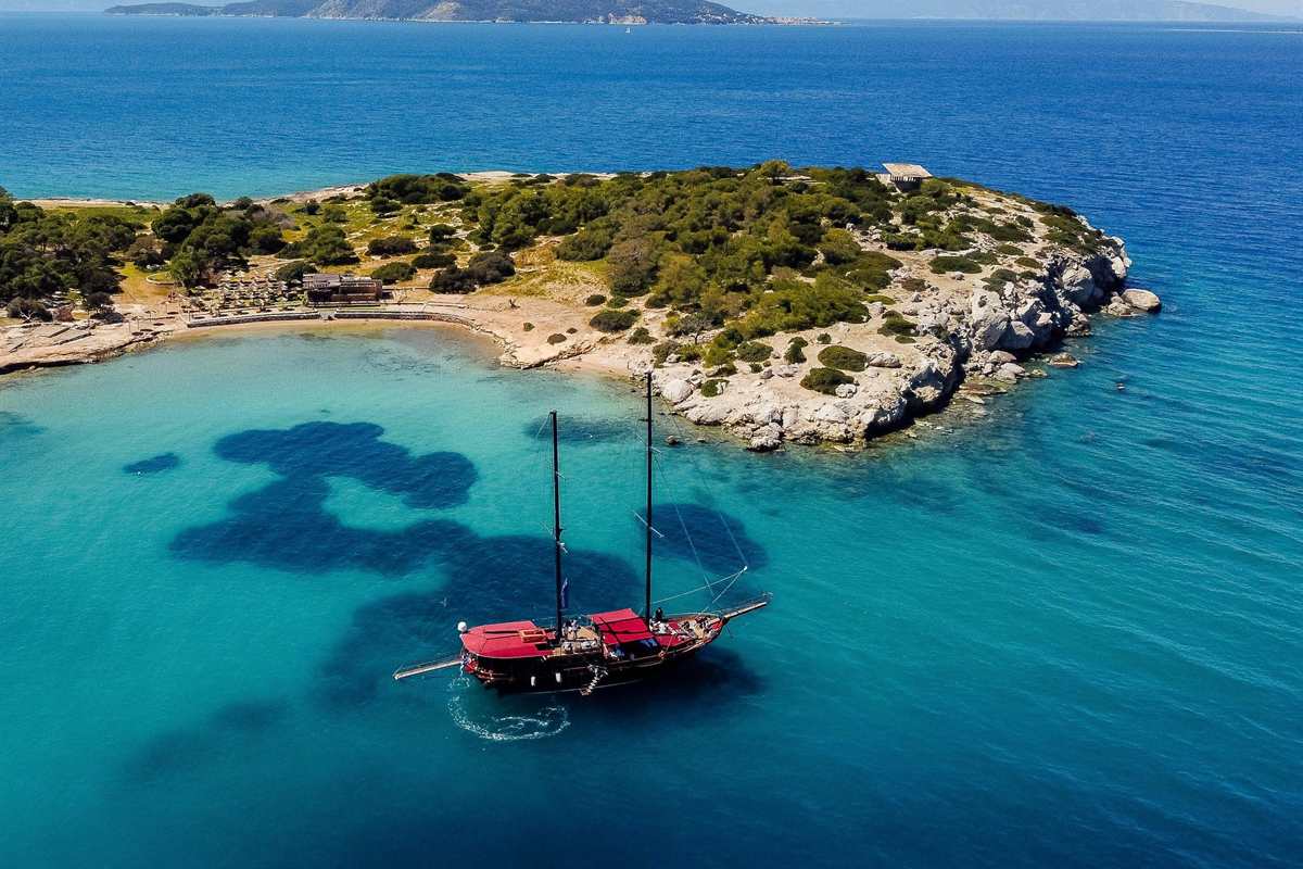 Agistri Moni Aegina cruise: red-roofed two-masted wooden gulet anchored in a clear turquoise bay beside a small pine-covered islet with a sandy cove and beach umbrellas, with the hills of another island on the horizon