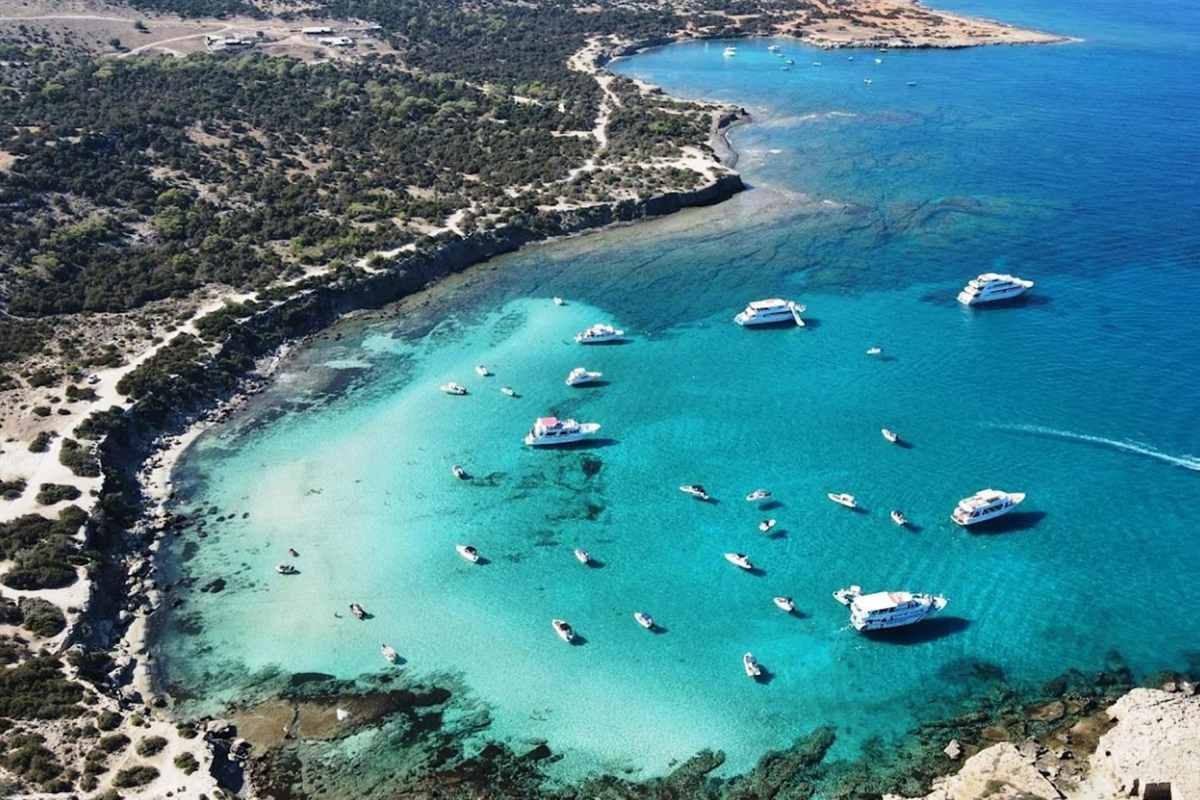 Akamas jeep safari: an aerial view of the Blue Lagoon bay near Latchi, with white cruise boats and small motorboats anchored on pale turquoise shallows, a dark green pine headland on the left and deep blue open sea on the right