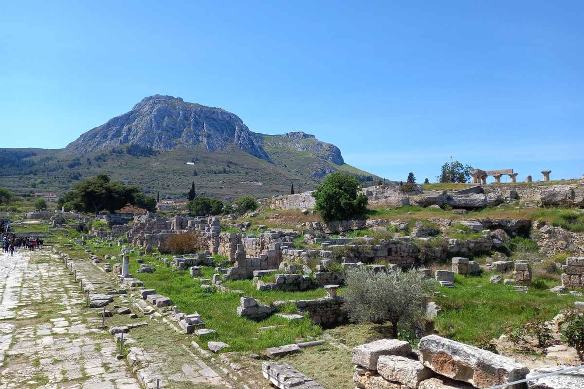 Ancient Corinth day trip from Athens: the ruined Agora of Ancient Corinth with a paved stone road on the left, green grass between low stone walls and column bases, the Temple of Apollo columns on the ridge at top right and the rocky peak of Acrocorinth behind under a clear blue sky