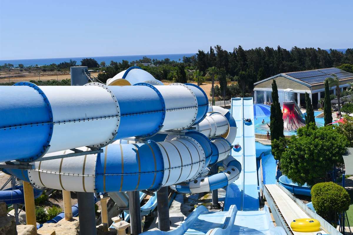 Aphrodite Waterpark tickets: a view from the top of the slides at the water park in Paphos, with wide blue and white slide tubes in the foreground, a long twin-lane slide, the red volcano of the leisure pool and the blue sea on the horizon under a clear sky