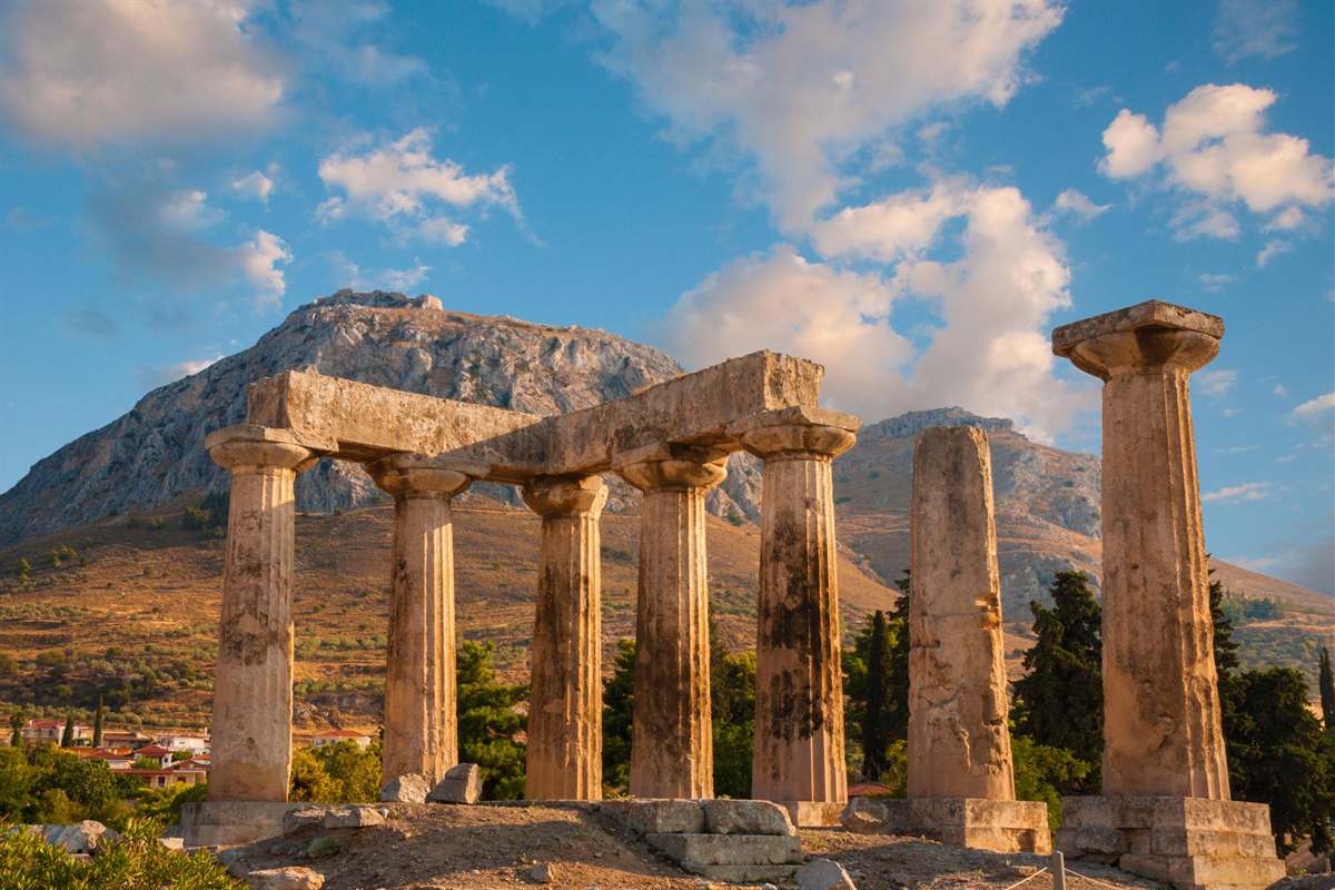 Apostle Paul tour Athens and Corinth: the Doric columns of the Temple of Apollo at Ancient Corinth glowing in warm light, with a stone lintel across the left columns and the grey rocky peak of Acrocorinth rising behind them under a blue sky with white clouds