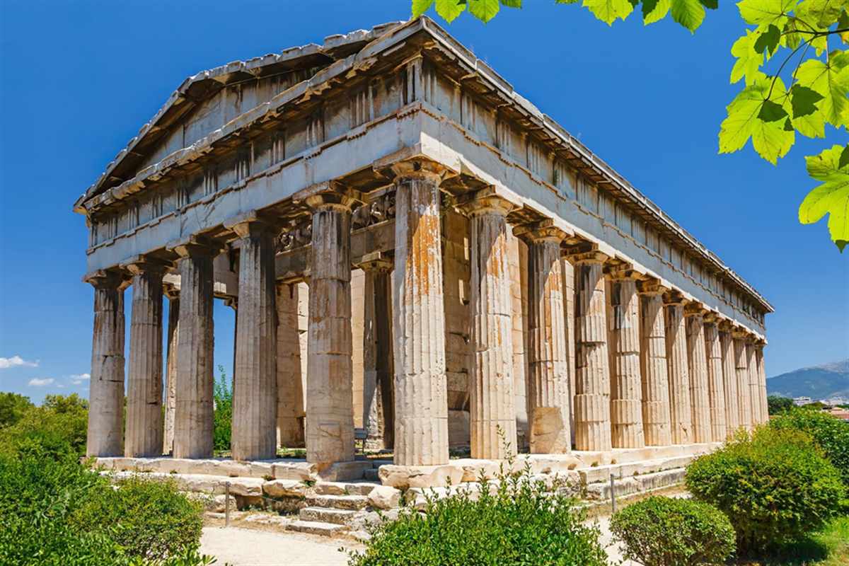 Apostle Paul tour Athens and Corinth: the Doric Temple of Hephaestus in the Ancient Agora of Athens seen from one corner, with tall fluted honey-coloured columns, a carved frieze, green shrubs around its base and a leafy branch at the top right under deep blue sky