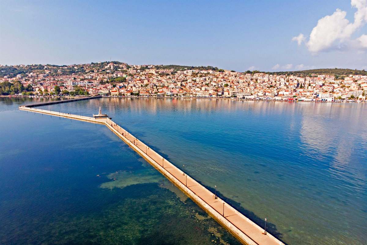 Argostoli boat trip: the long De Bosset Bridge stretching across the calm blue bay of Argostoli, with its white obelisk at the bend and the red-roofed houses of the town climbing the green hillside behind under a clear sky