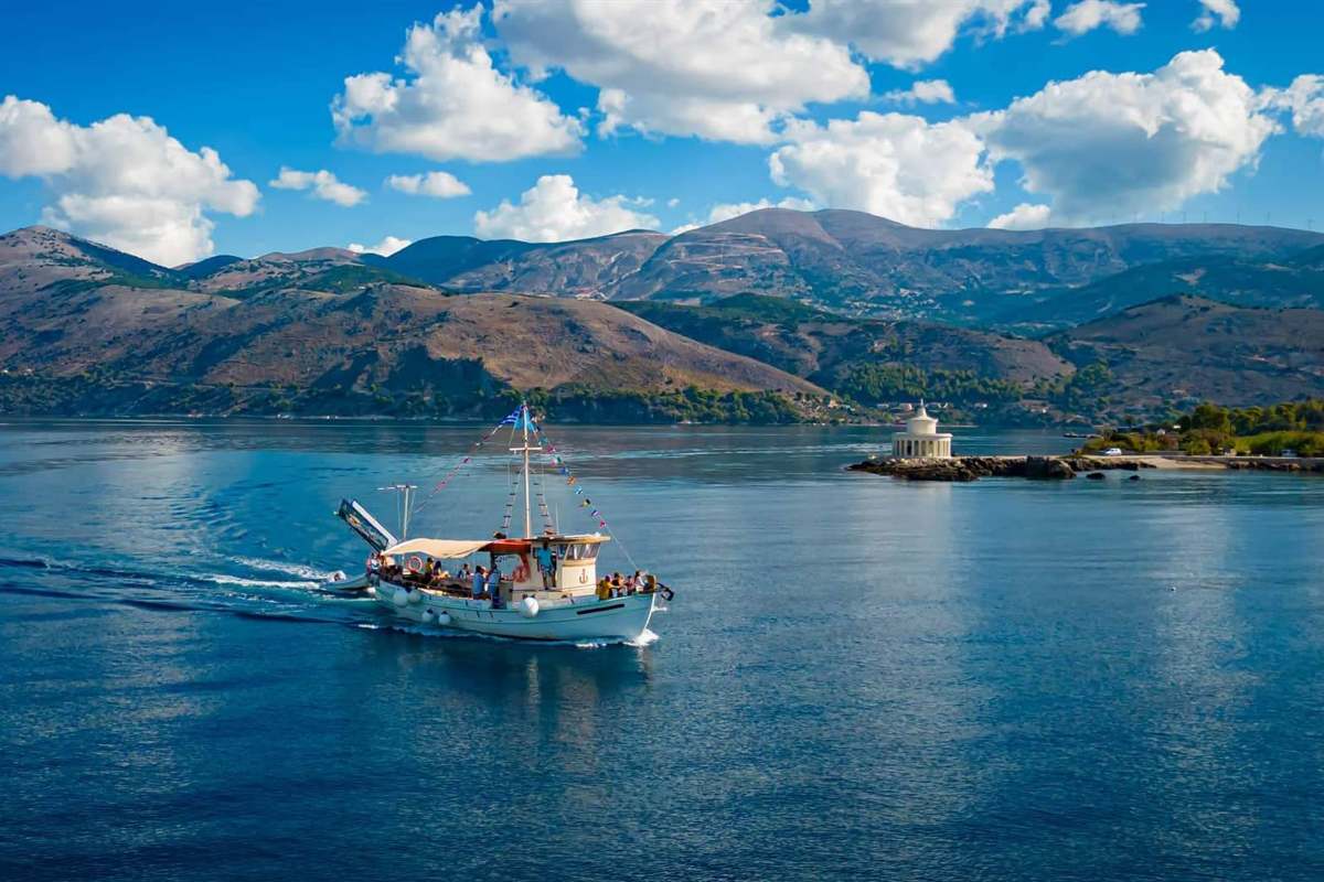Argostoli boat trip: a white traditional Greek boat with a canvas awning and a string of bunting flags cruising across the deep blue Argostoli gulf towards the round Lighthouse of Saint Theodore, with green mountains and white clouds behind