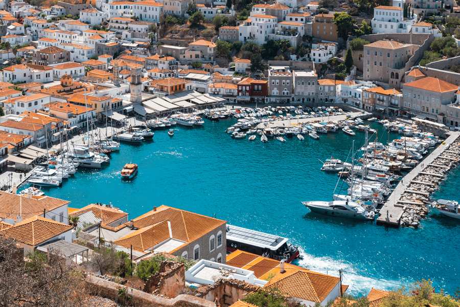 Arrange your own tour of Greece: aerial view of a harbour town with orange tiled roofs, white houses and moored boats beside deep blue water