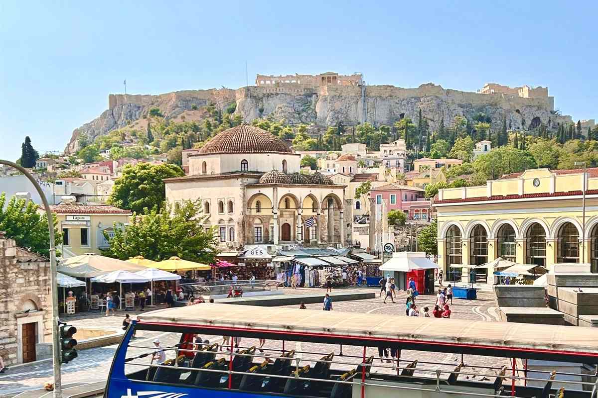 Athens hop on hop off bus: the open upper deck of a blue double-decker bus with empty seats in the foreground above Monastiraki Square, with the domed Tzistarakis Mosque, cafe umbrellas and market stalls, and the walls and ruins of the Acropolis rising on the rocky hill behind under a blue sky