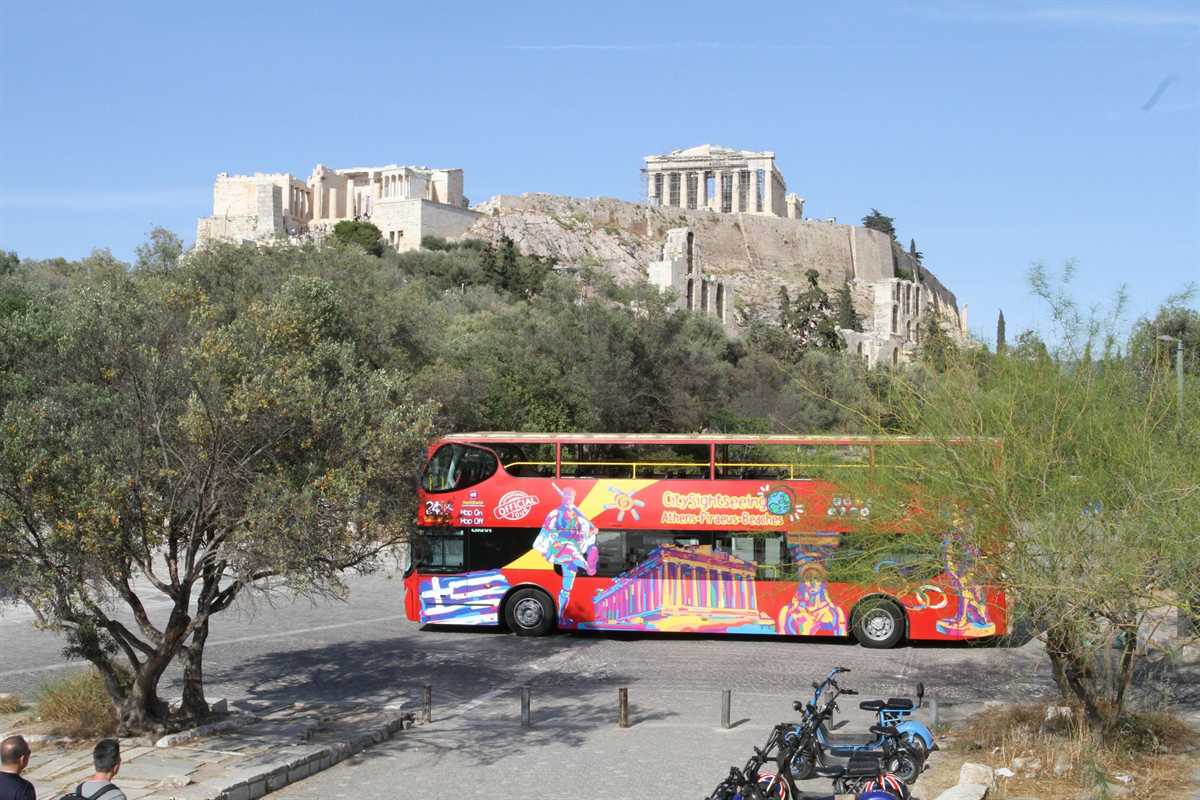 Athens hop on hop off bus: a red open-top double-decker bus with a colourful Greek flag and Parthenon design on its side, parked on a paved square behind an olive tree, with the Propylaea and the Parthenon on the rocky Acropolis hill above and the arched stone wall of the Odeon of Herodes Atticus under a clear blue sky