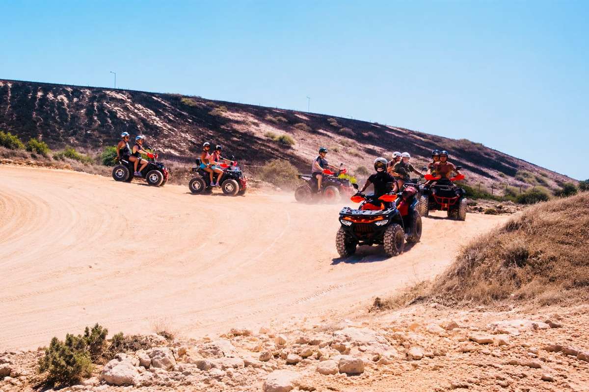Ayia Napa quad safari: a group of quad bikes with helmeted riders sweeping around a wide curve of sandy dirt track below a dry hillside, with dust rising behind them, rocks and low scrub in the foreground and a clear blue sky above