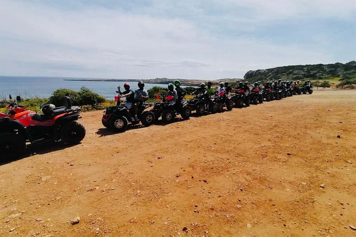 Ayia Napa quad safari: a long line of quad bikes with helmeted riders parked side by side on a bare red-brown dirt clearing at the edge of a coastal headland, with green scrub and yellow wildflowers beside them, calm blue sea and a low rocky cape behind under a pale cloudy sky