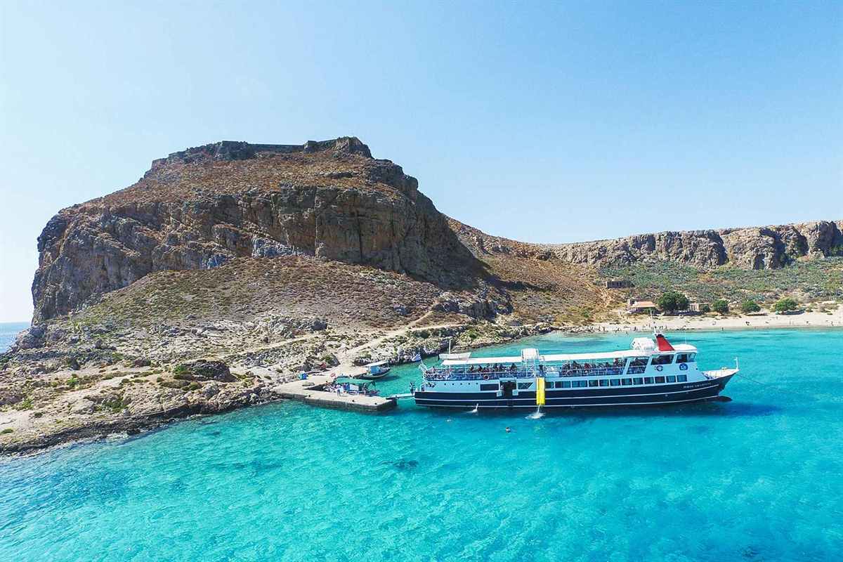 Balos Gramvousa boat trip from Chania: a white and navy cruise ship moored at the small stone pier of Imeri Gramvousa in clear turquoise water, below the rocky hill topped by the Venetian fortress