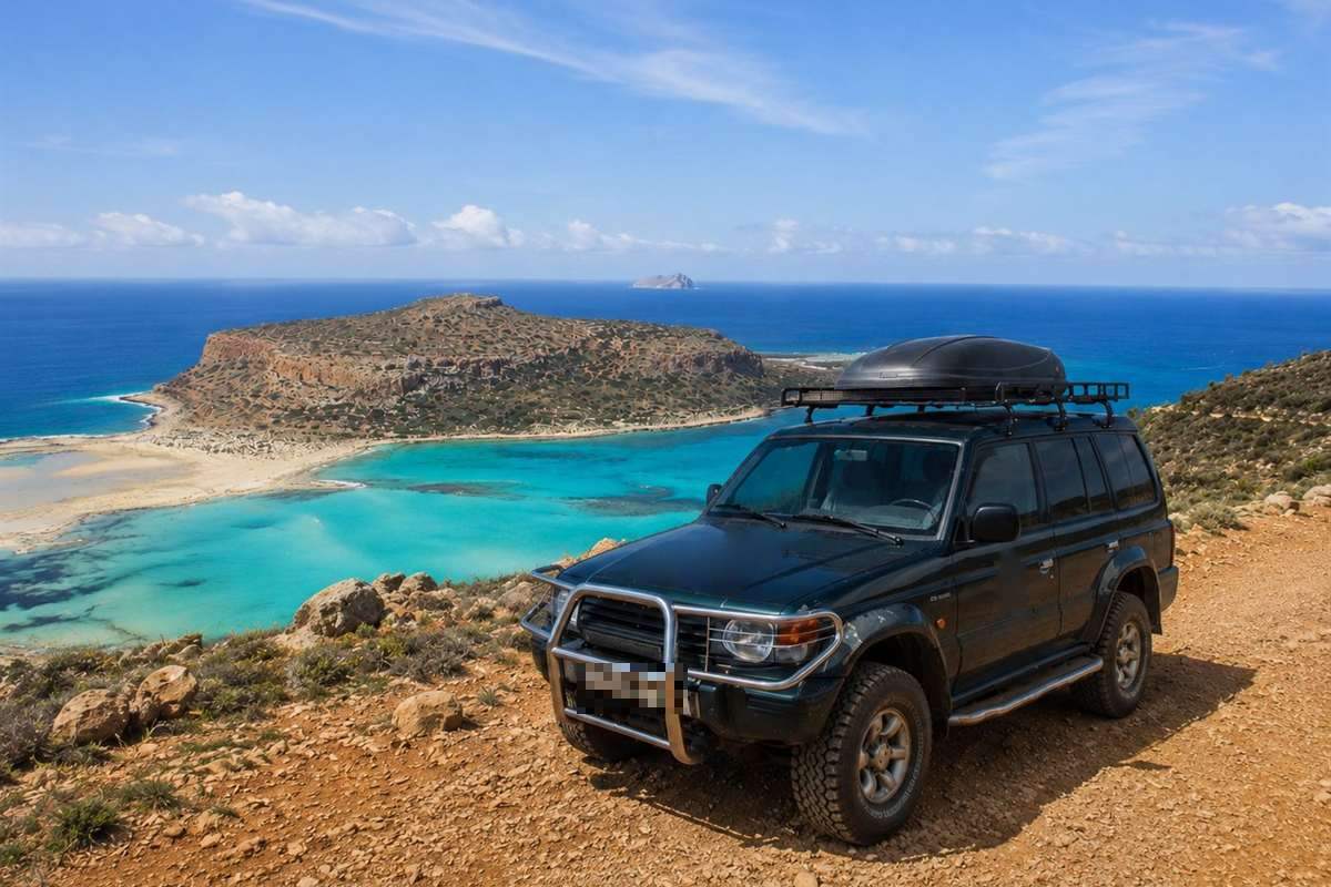 Balos jeep safari: a dark green 4x4 with a roof rack and a black roof box parked on an orange dirt track on a hilltop, with the turquoise Balos lagoon, the flat-topped rocky headland of Cape Tigani and deep blue sea below under a bright blue sky with thin cloud streaks