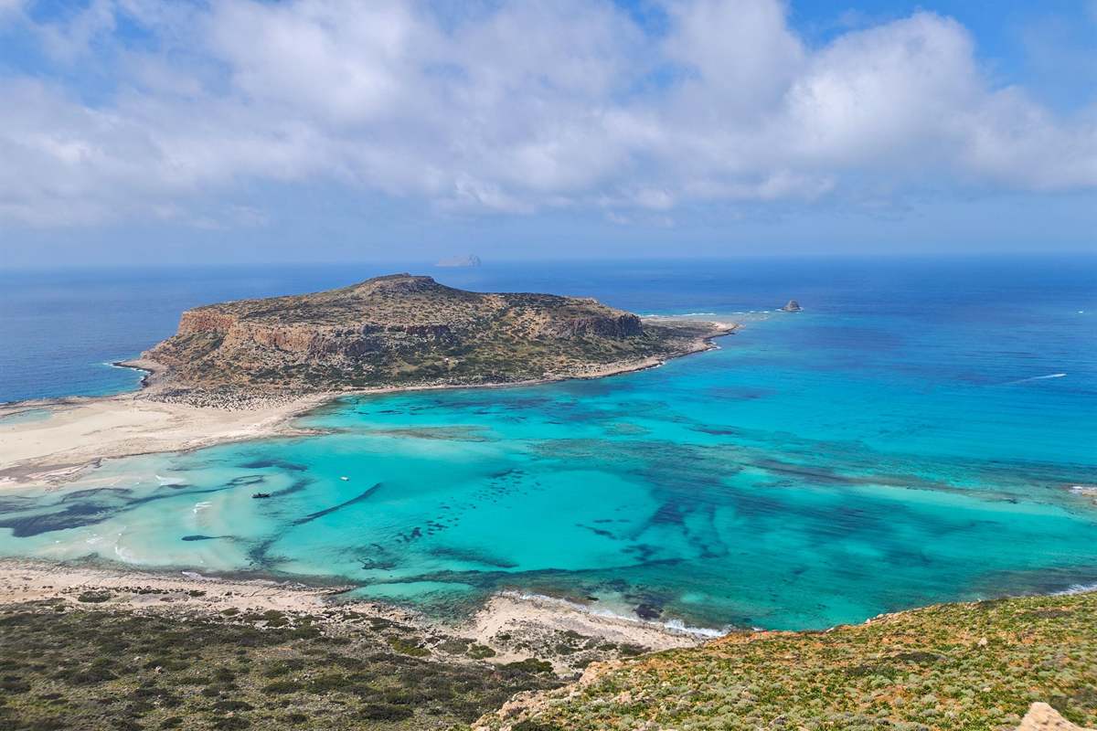 Balos jeep safari: the turquoise Balos lagoon seen from the hilltop above the dirt track, with pale sand bars, the flat-topped rocky headland of Cape Tigani, deep blue open sea and a sky of large white clouds