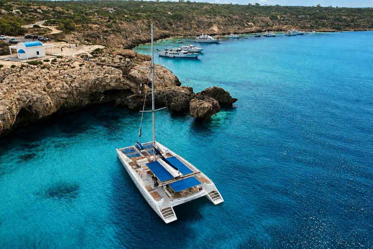 Blue Lagoon cruise Ayia Napa: a white sailing catamaran with blue sun awnings anchored in clear turquoise water beside a rocky headland with a small white building with a blue roof, with other cruise boats moored in the bay beyond under a blue sky