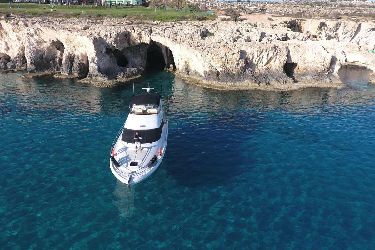 Blue Lagoon cruise Ayia Napa: a small white motor yacht anchored in clear blue-green water below pale limestone cliffs with dark sea cave openings, with grass and low buildings along the clifftop above