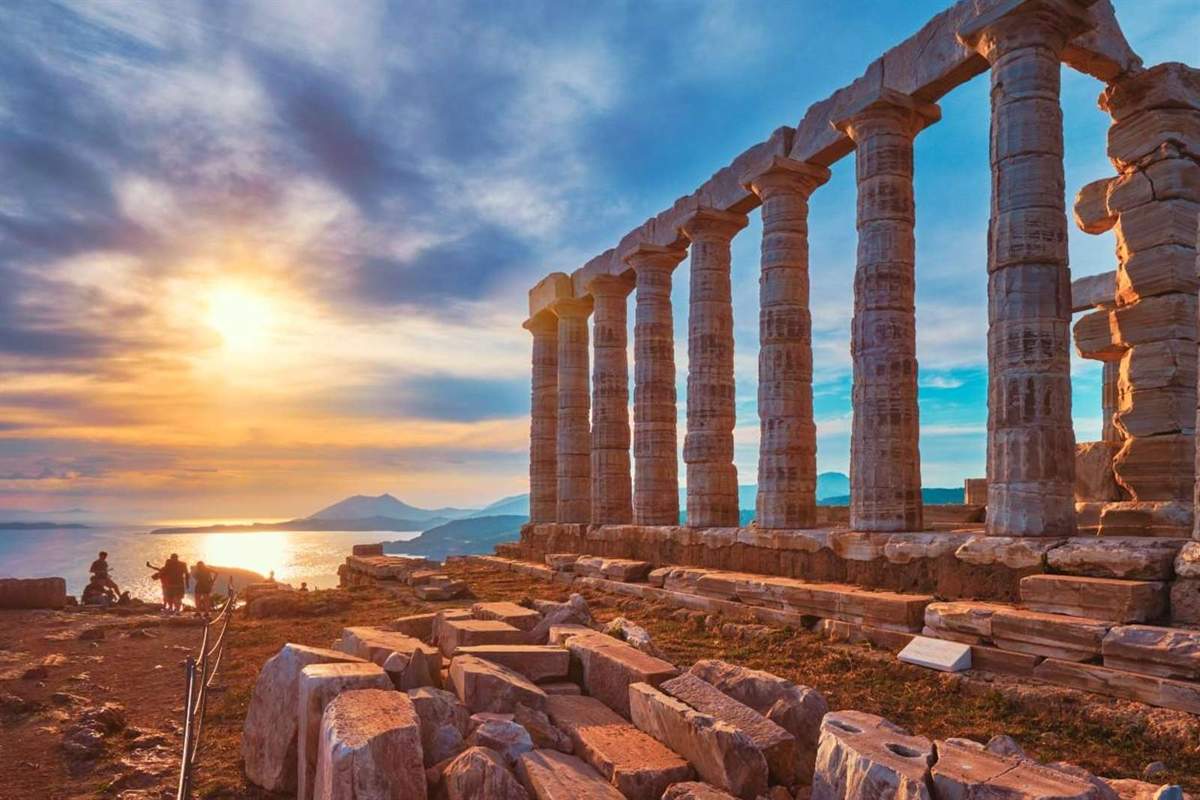 Cape Sounion sunset tour from Athens: golden sun sinking over the Aegean beside the tall columns of the Temple of Poseidon, with visitors watching from the cliff edge and fallen marble blocks in the foreground