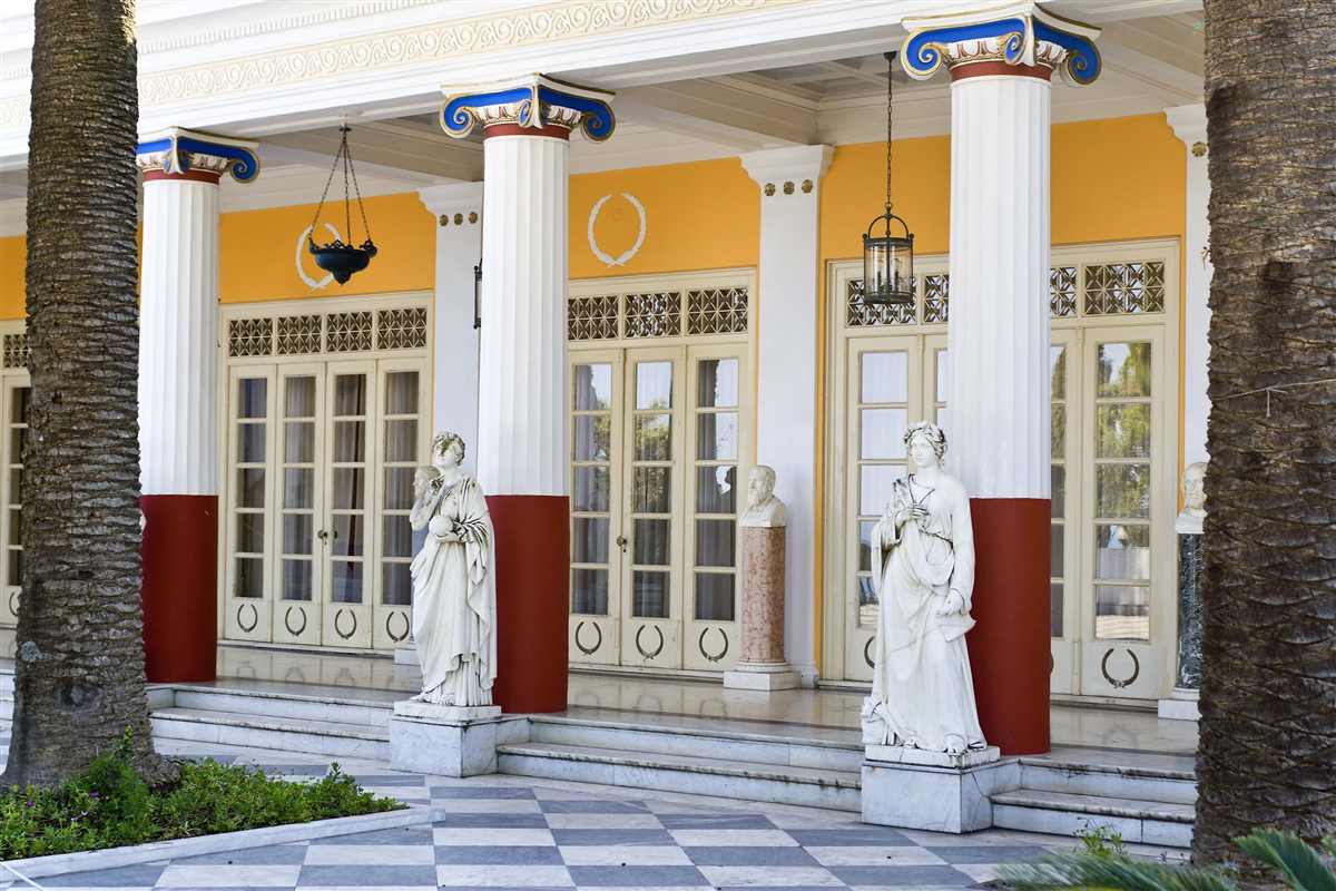 Corfu hop on hop off bus: the colonnade of the Achilleion Palace with white fluted columns, blue and gold capitals, red column bases, yellow walls and two white marble statues on a chequered marble terrace between palm trunks