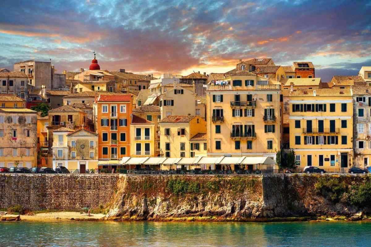 Corfu hop on hop off bus: pastel yellow and orange townhouses of Corfu Old Town with a red church dome and cafe awnings above the old sea wall and turquoise water, under a pink and orange sunset sky