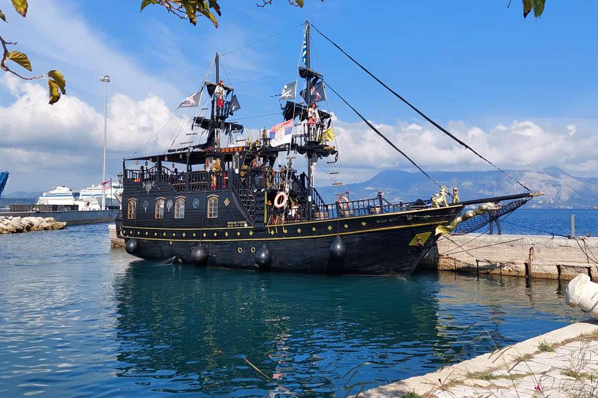 Corfu pirate ship: a black wooden pirate-style ship with two masts, black flags and gold trim moored at a stone quay beside calm blue water, with white cruise ships and a mountain coast behind and green leaves framing the sky